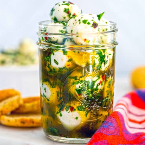 Marinated mozzarella balls with lots of herbs in a mason jar with crostini in background.