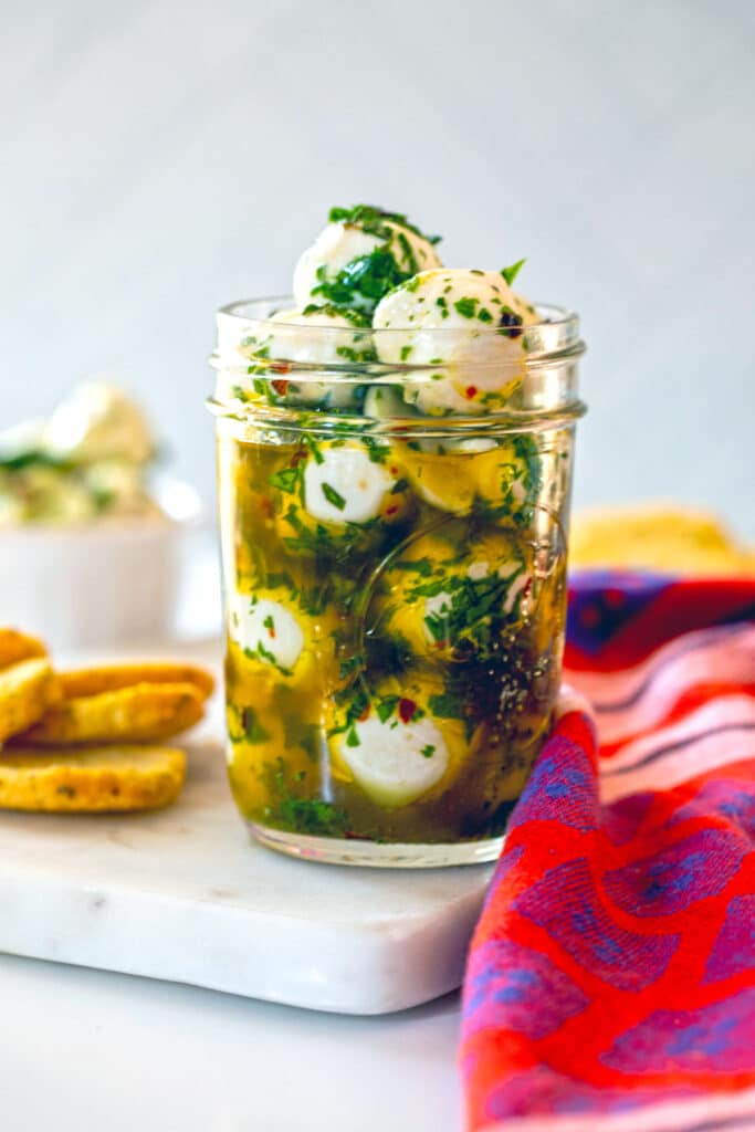 Jar filled with mozzarella balls in olive oil with crostini and more mozzarella pearls on the side.