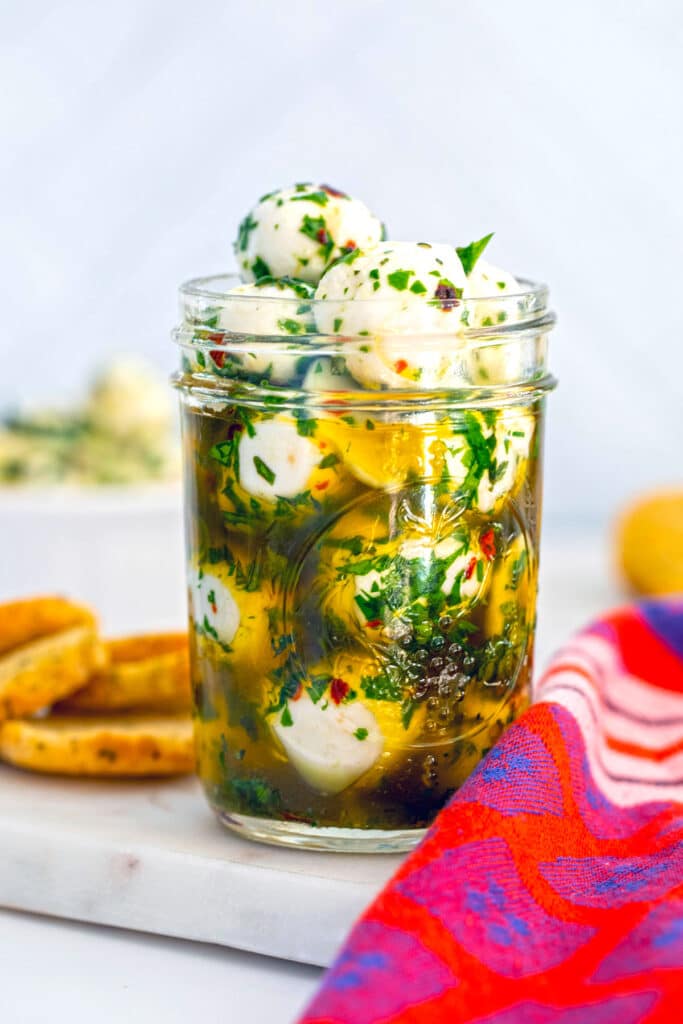 Mozzarella balls in a jar with oil and herbs with crostini in the background.