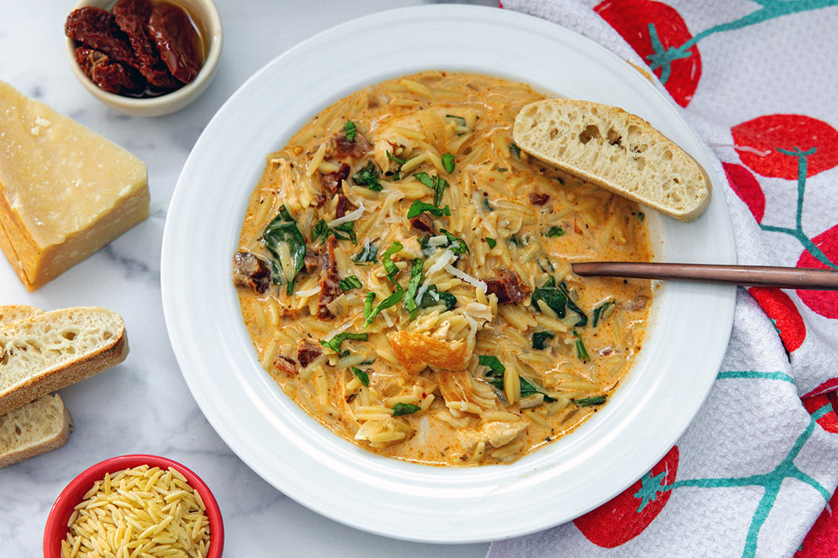 Landscape head-on view of soup in bowl with bread, sundried tomatoes, parmesan, and orzo on the side.