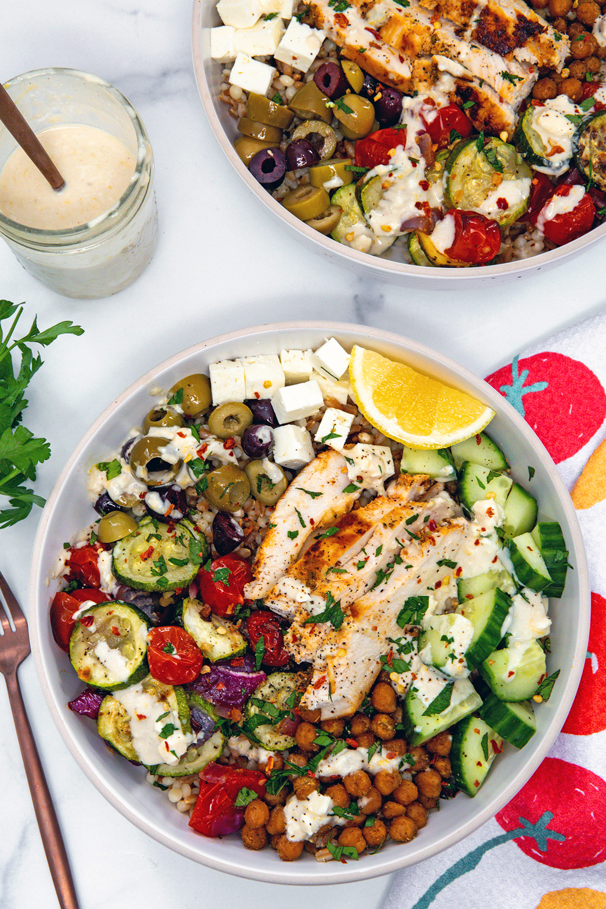 Overhead view of a Mediterranean grain bowl with chicken, zucchini, tomatoes, cucumbers, red onion, feta, and olives with second bowl in background, and jar of dressing.