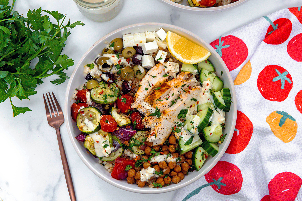 Overhead landscape view of a Mediterranean grain bowl with chicken and veggies with fork and bundle of parsley in background.