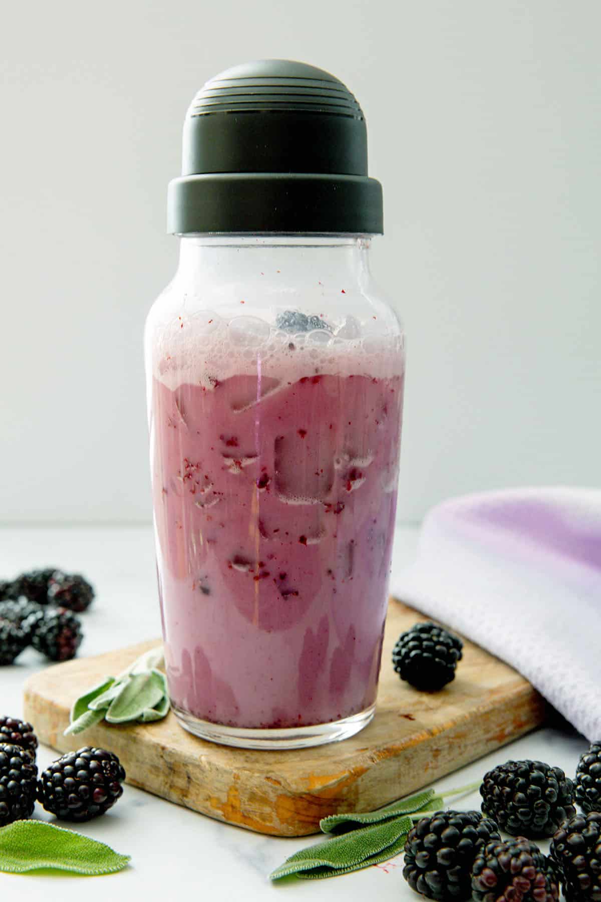 Midnight Drink in a clear shaker with blackberries and sage leaves all around.