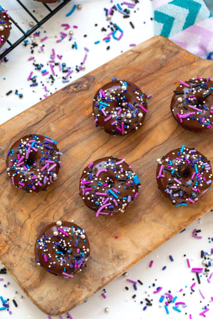 Mini chocolate donuts on a wooden cutting board with sprinkles all around.