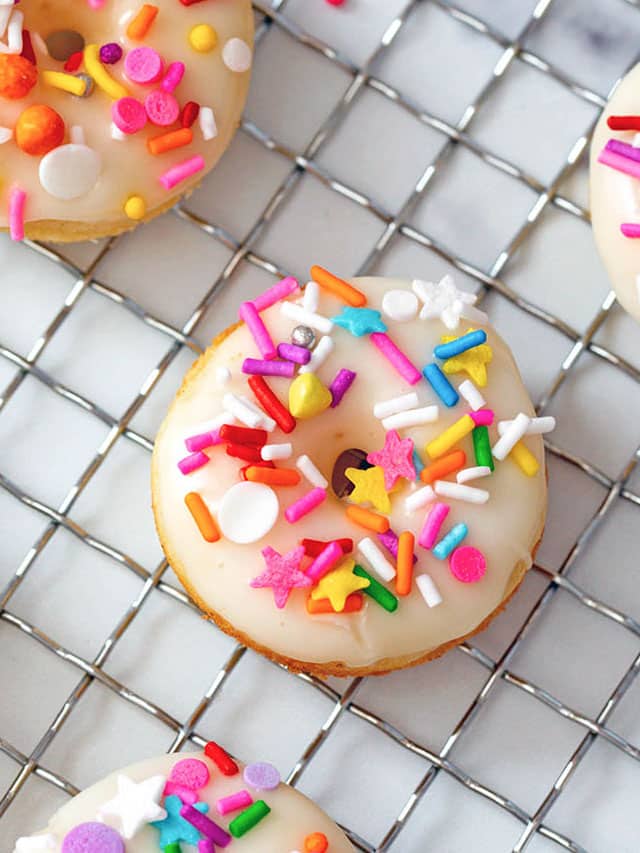 Mini donut with vanilla icing and rainbow sprinkles on cooling rack.