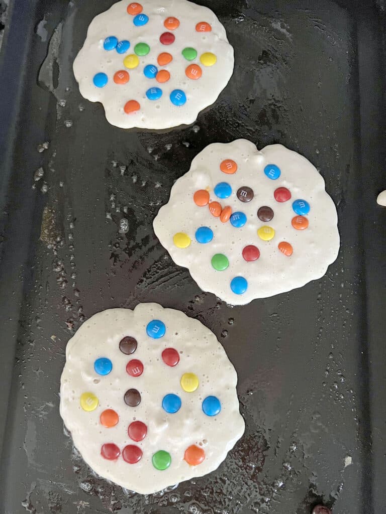 Pancake batter with mini M&Ms cooking on a griddle.