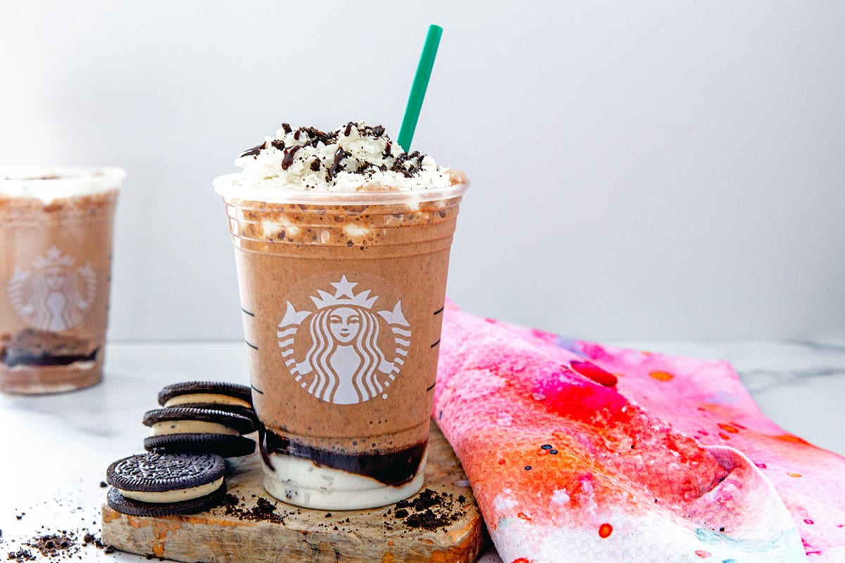 Landscape head-on view of a Mocha Cookies Crumble Frappuccino with green straw with second drink in background and cookies around.