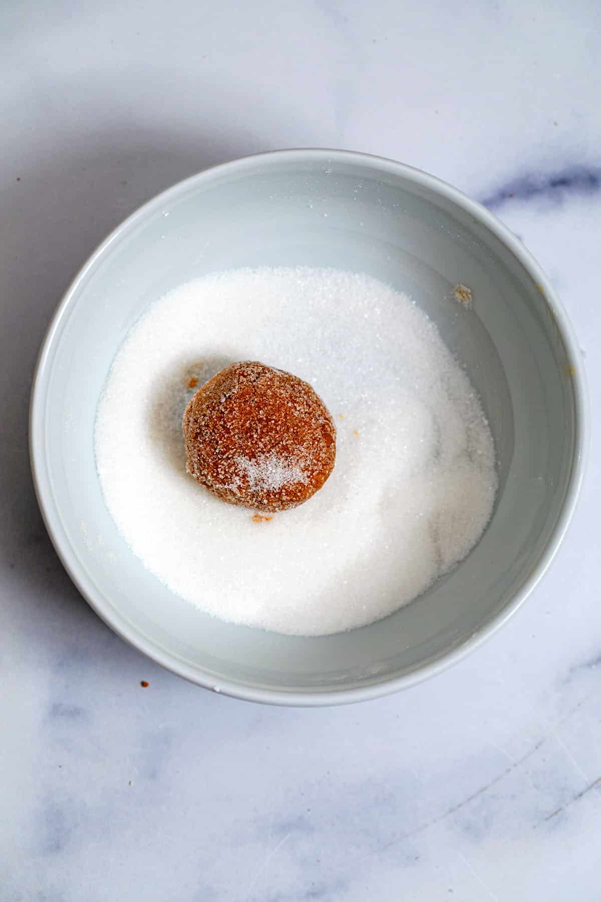 Cookie dough ball being rolled in granulated sugar.