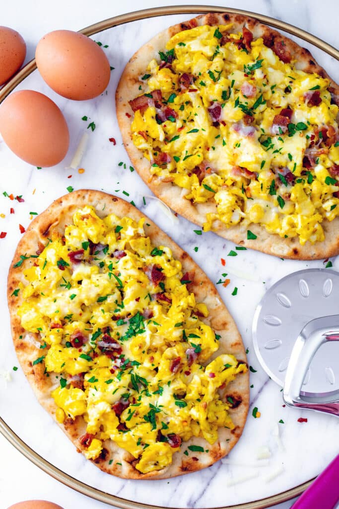 Two naan breakfast pizzas on marble tray with pizza cutter and whole eggs.