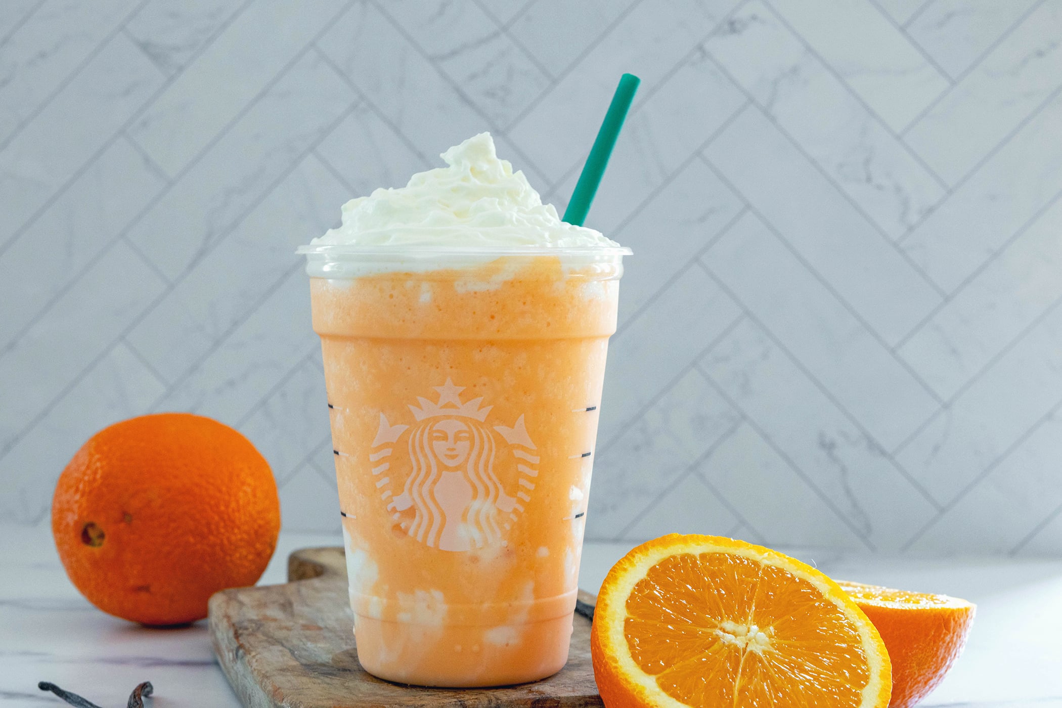 Landscape head-on view of an Orange Cream Frappuccino with whipped cream and green straw with oranges all around.