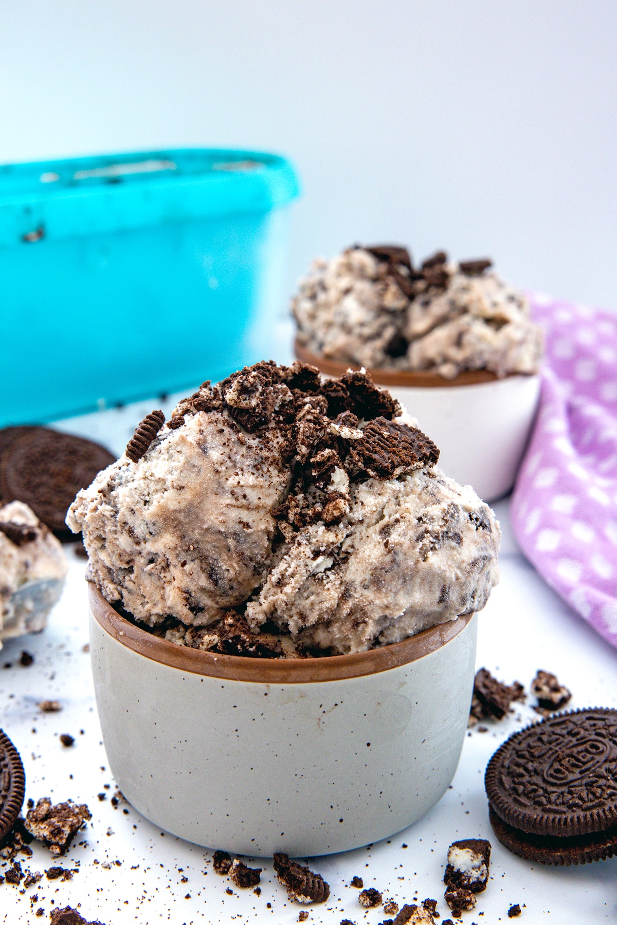 Head-on view of a bowl of Oreo ice cream with second bowl and container in background and Oreo cookies all around.