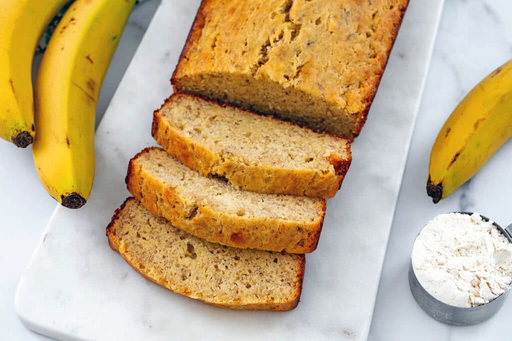 Overhead view of a loaf of banana bread with slices cut, fresh bananas surrounding it, and cup of pancake mix on the side.