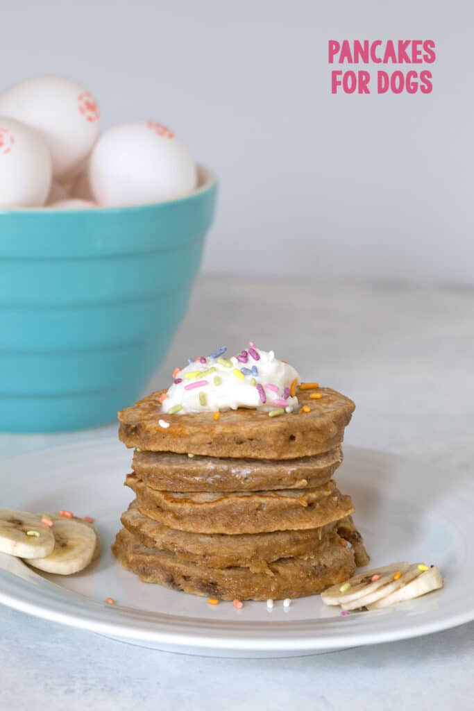 Stack of egg, apple, and banana pancakes for dogs with whipped cream and sprinkles and bowl of eggs in the background with recipe title at top.