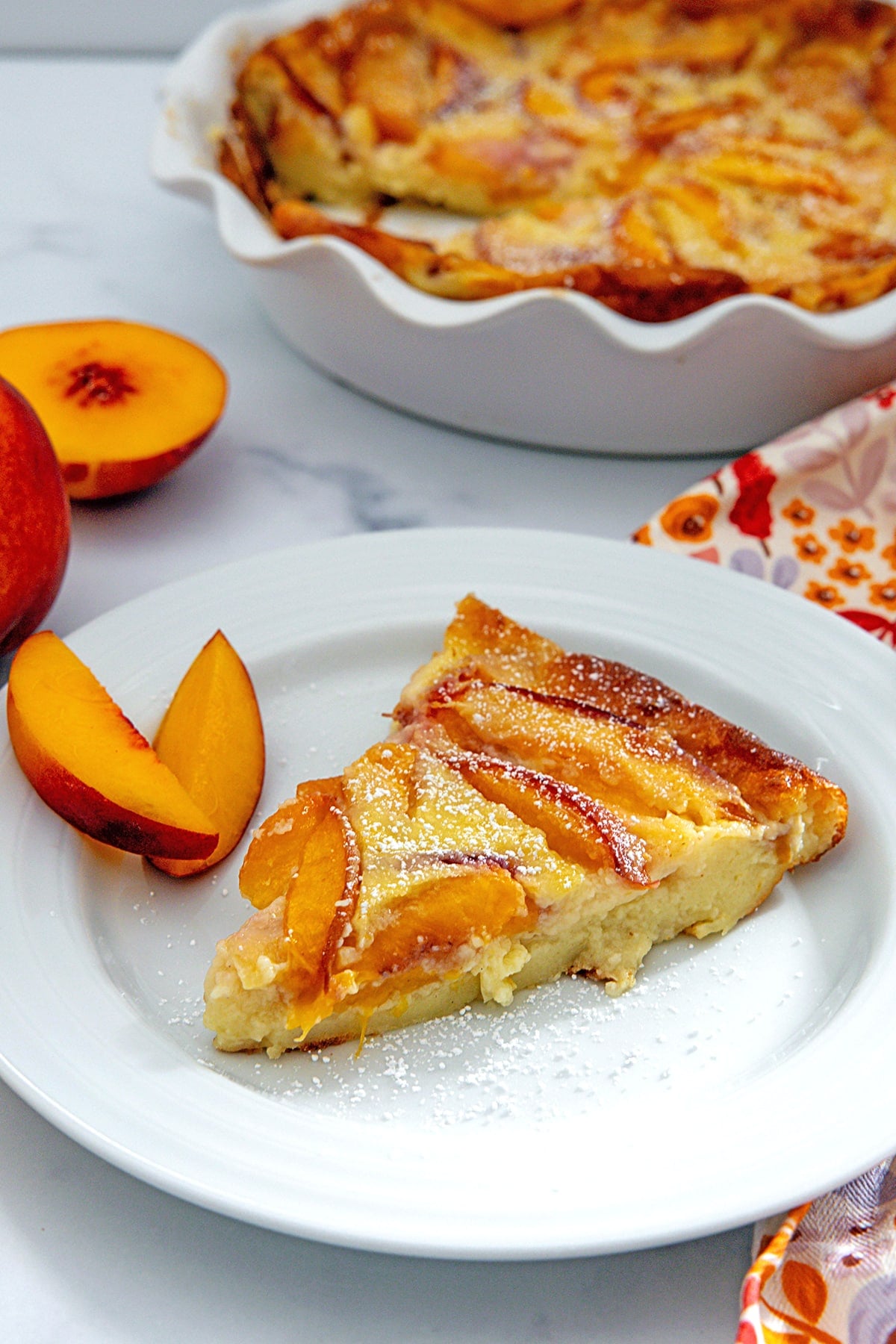 Head-on view of a slice of peach clafoutis with whole clafoutis in the background and peach slices all around.