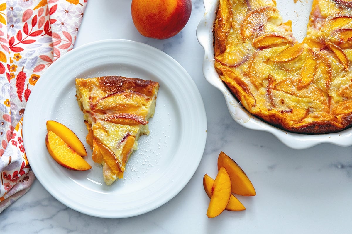 Overhead landscape view of a peach clafoutis in a pie dish with a slice on a plate to the side and peach slices all around.