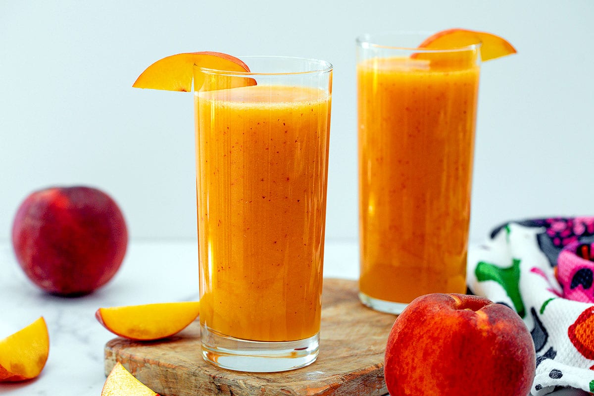 Head-on landscape view of two tall glasses of peach juice with peaches all around.