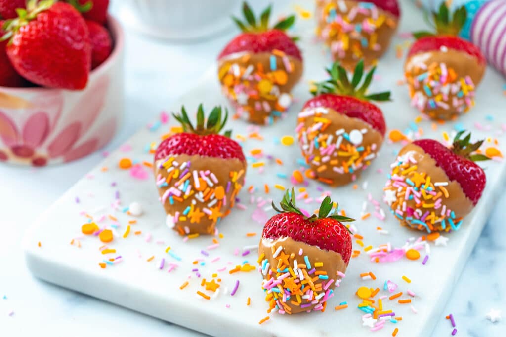 Landscape view of multiple peanut butter strawberries with sprinkles on a marble platter with bowl of fresh strawberries in background.