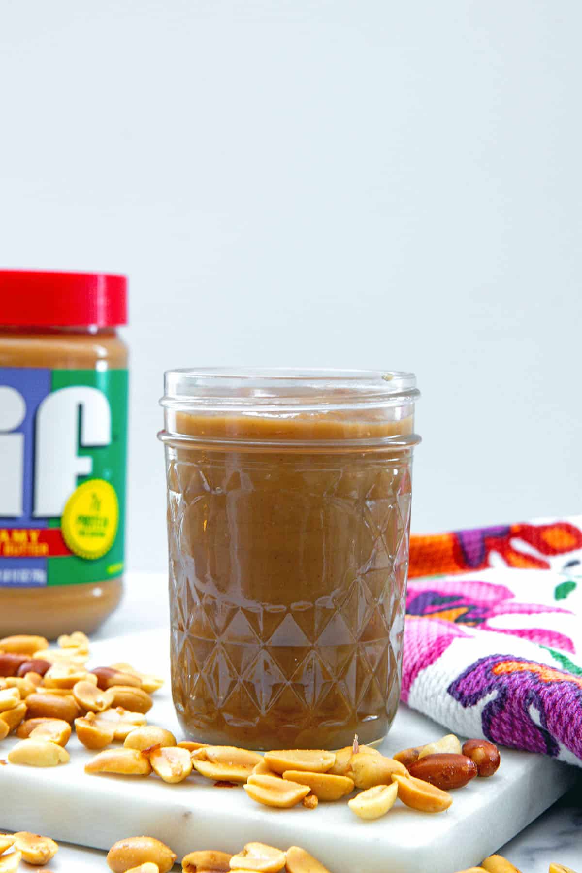 Head-on view of a jar of peanut butter syrup with Jif peanut butter container in background and peanuts all around.