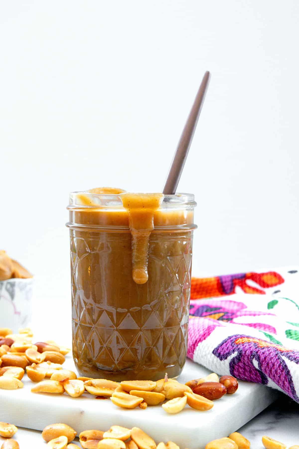 Head-on view of a jar of peanut butter syrup with spoon with syrup dripping out of jar and peanuts all around.