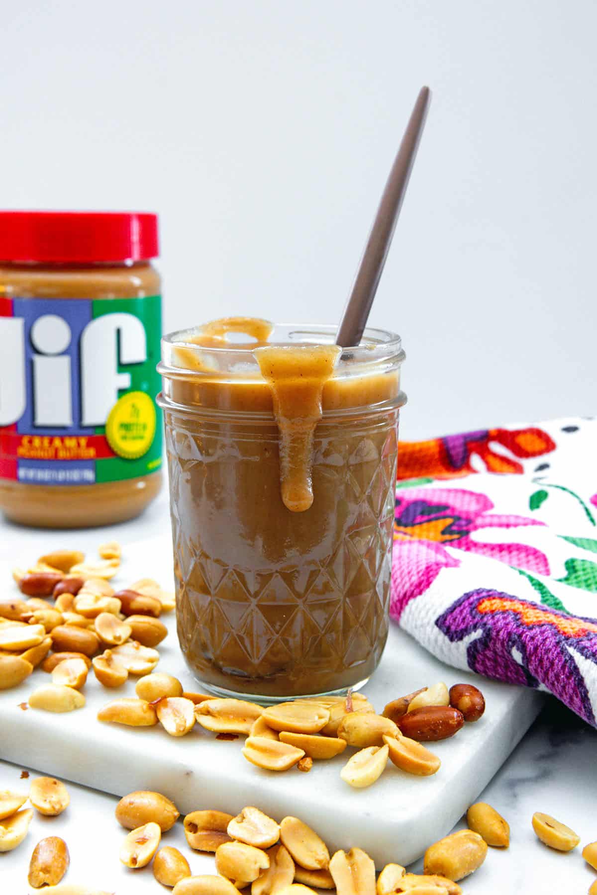 Peanut butter syrup drizzling over the side of a jar with peanuts all around and container of Jif peanut butter in background.