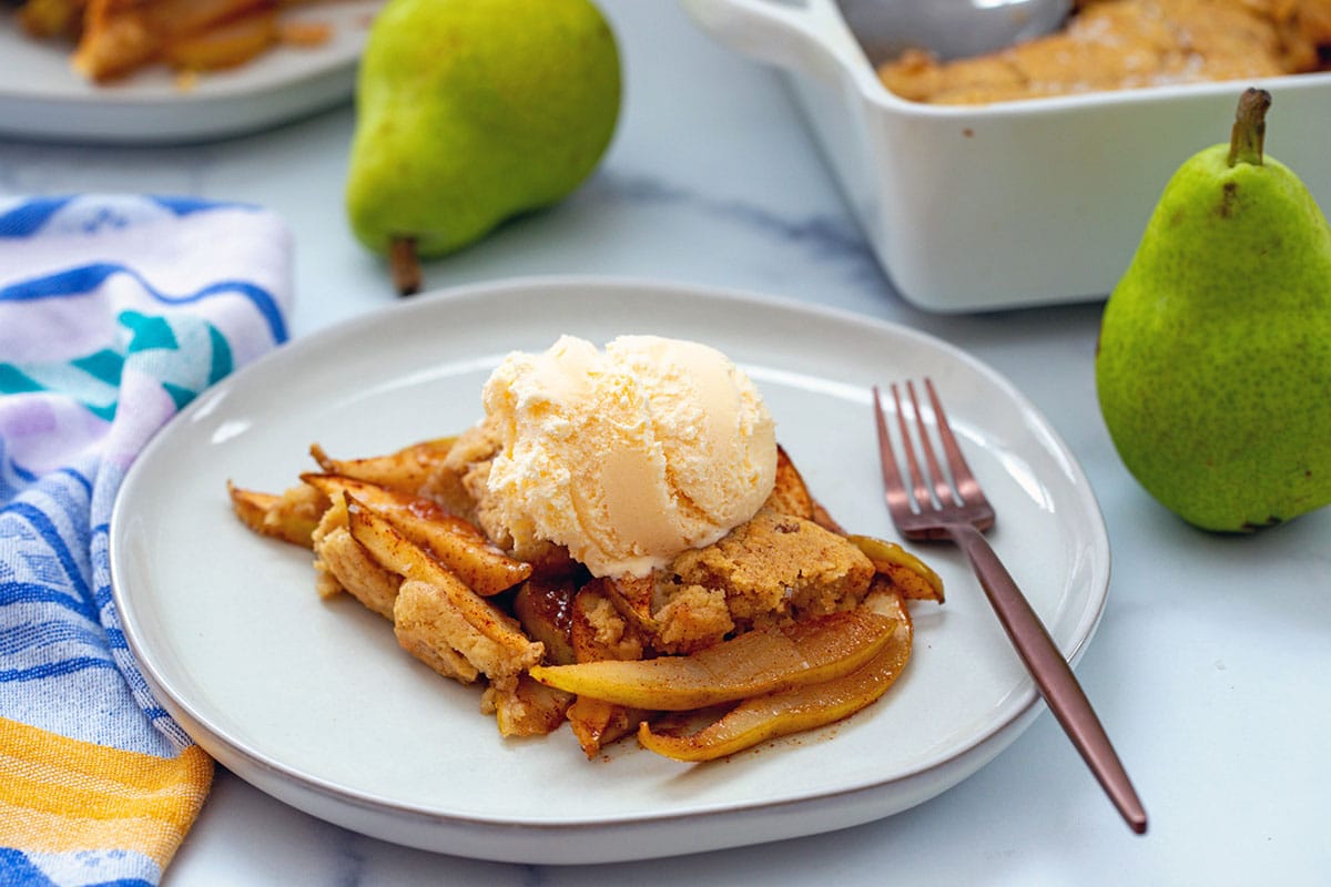 Landscape head-on view of pear cobbler topped with vanilla ice cream on plate with pears and baking dish in background.
