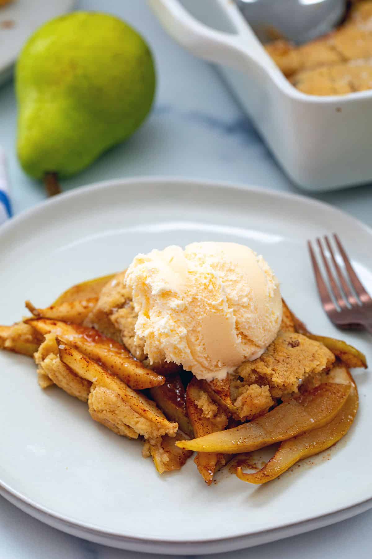 Pear cobbler on a plate topped with vanilla ice cream with pear and baking dish in background.