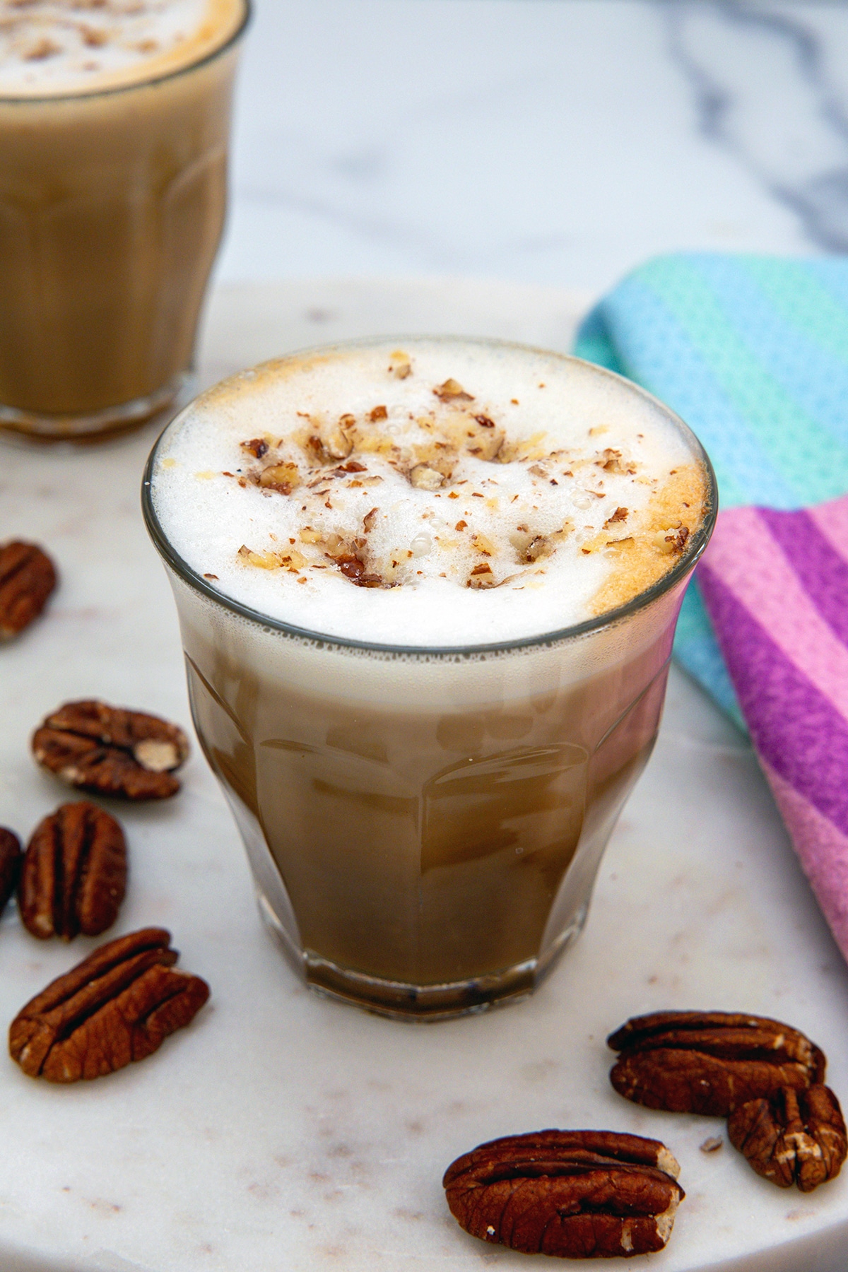 Overhead view of foam on top of a Pecan Oatmilk Cortado with second glass in background and pecans all around.