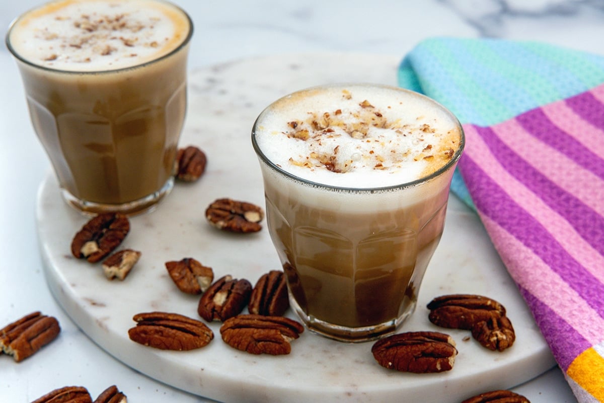Landscape overhead view of two glasses of Pecan Oatmilk Cortado with froth on top and with pecans all around.