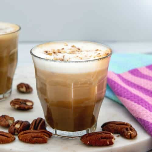Closeup view of a Pecan Oatmilk Cortado in a glass with pecans all around.