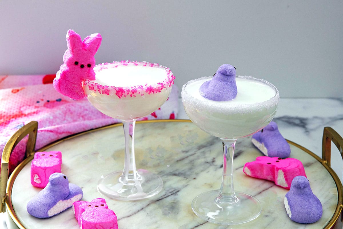Landscape view of two Peeps cocktails in coupe glasses on a marble surface with pink and purple Peeps all around.