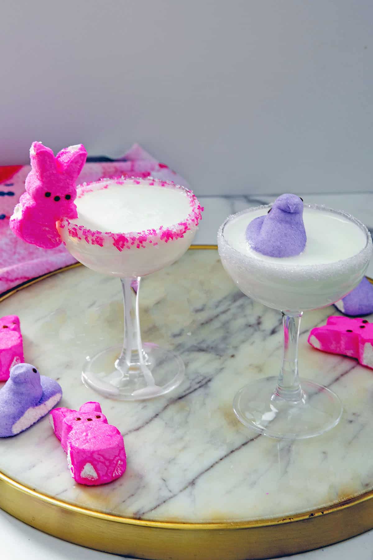 Two white cocktails in couple glasses, one with pink sugar rim and bunny Peep and one with purple sugar rim and chick Peep.
