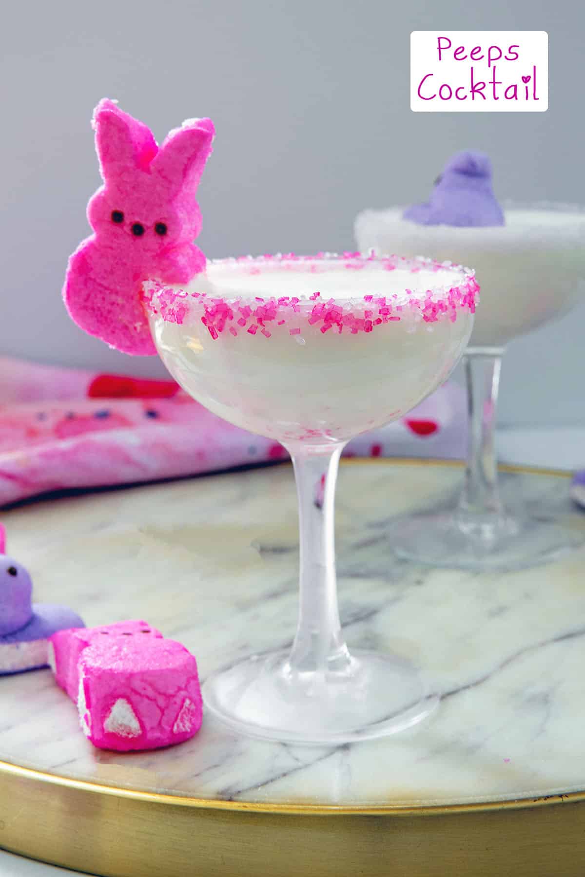 Head-on view of a white cocktail in a coupe glass with pink bunny Peeps on the side with second cocktail in background and recipe title at top.