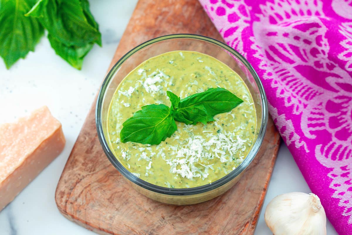 Landscape overhead view of small bowl of pesto cream sauce with fresh basil,  block of parmesan cheese, and garlic clove.