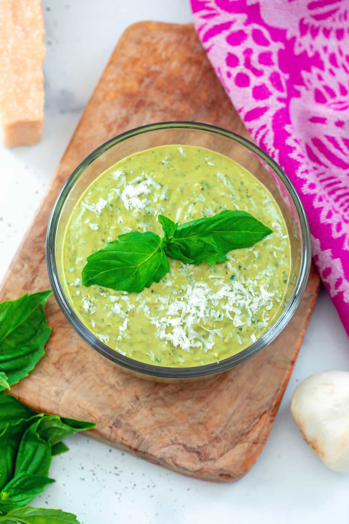 Overhead view of pesto cream sauce in bowl with fresh basil, garlic clove, and block of parmesan in background.