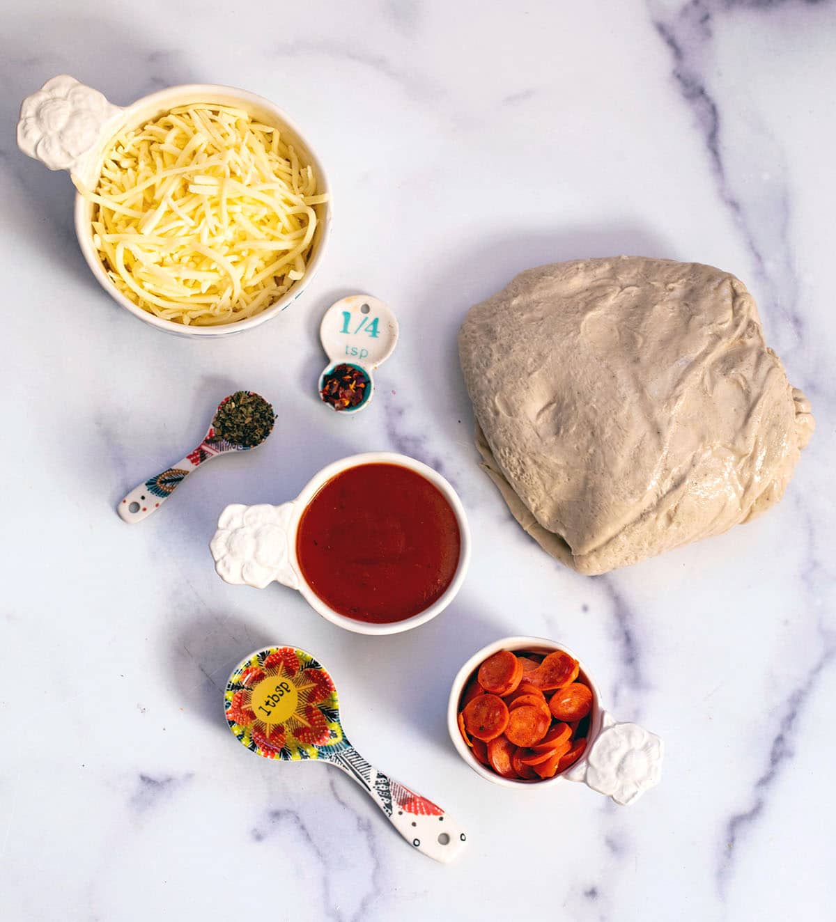 Overhead view of ingredients needed for pizza cupcakes, including: pizza dough, shredded cheese, pizza sauce, mini pepperoni, olive oil, dried basil, and red pepper flakes.