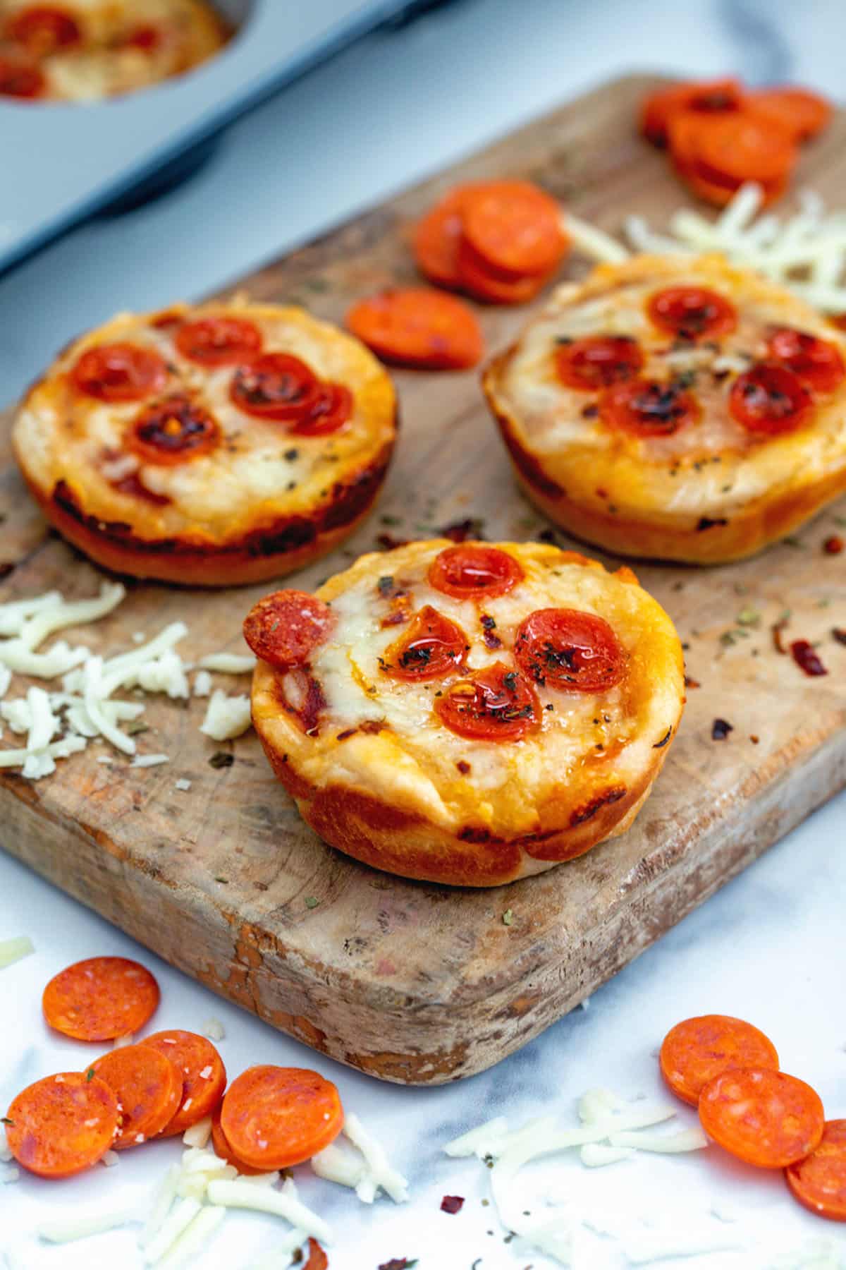 Head-on view of pizza cupcakes on wooden board with mini pepperoni and shredded cheese all around.