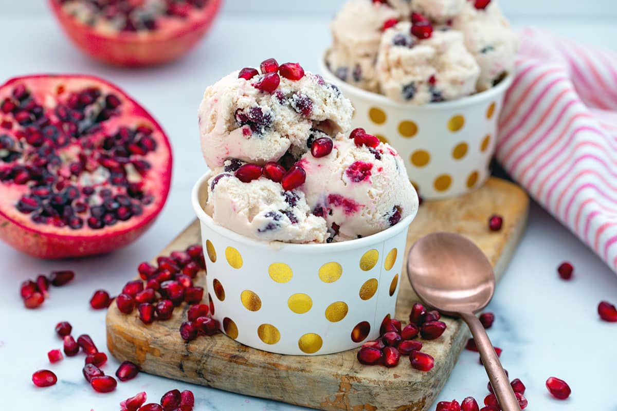 Landscape head-on view of two bowls of pomegranate ice cream with fruit halves in background and arils all around.