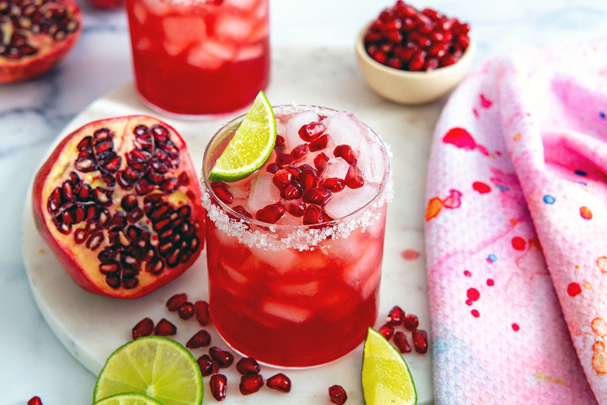 Landscape overhead view of a pomegranate margarita with half a pomegranate and arils and lime wedges all around.