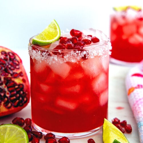 Closeup view of a pomegranate margarita with limes and arils all around.