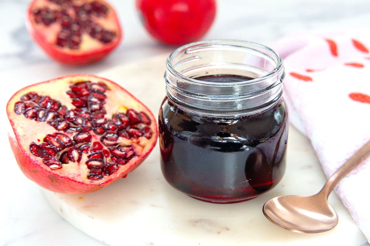 Head-on view of a small jar of pomegranate syrup with spoon and pomegranate half on the side.
