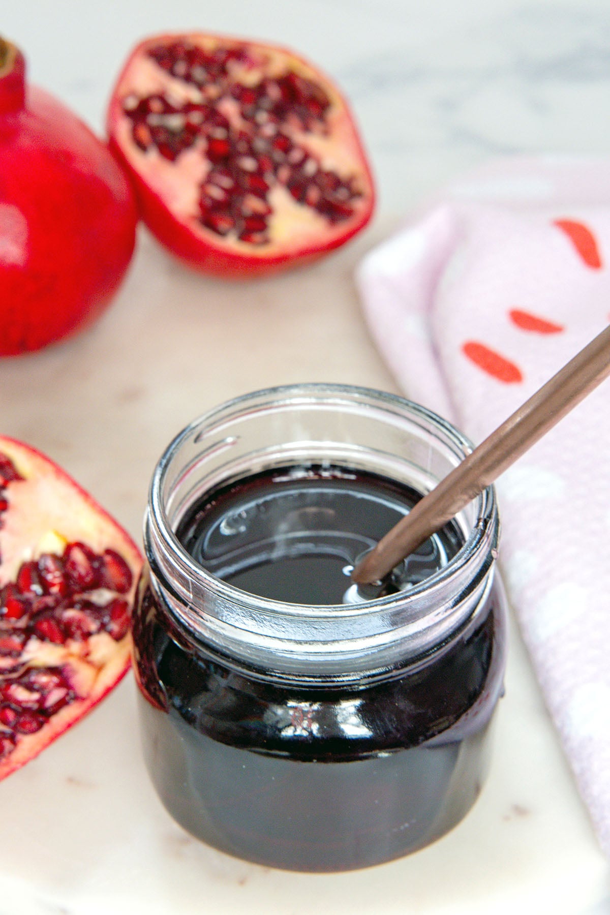 Overhead view of small jar of pomegranate syrup with spoon in it with pomegranate halves all around.