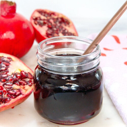 Closeup view of a small jar of pomegranate syrup with pomegranate halves.