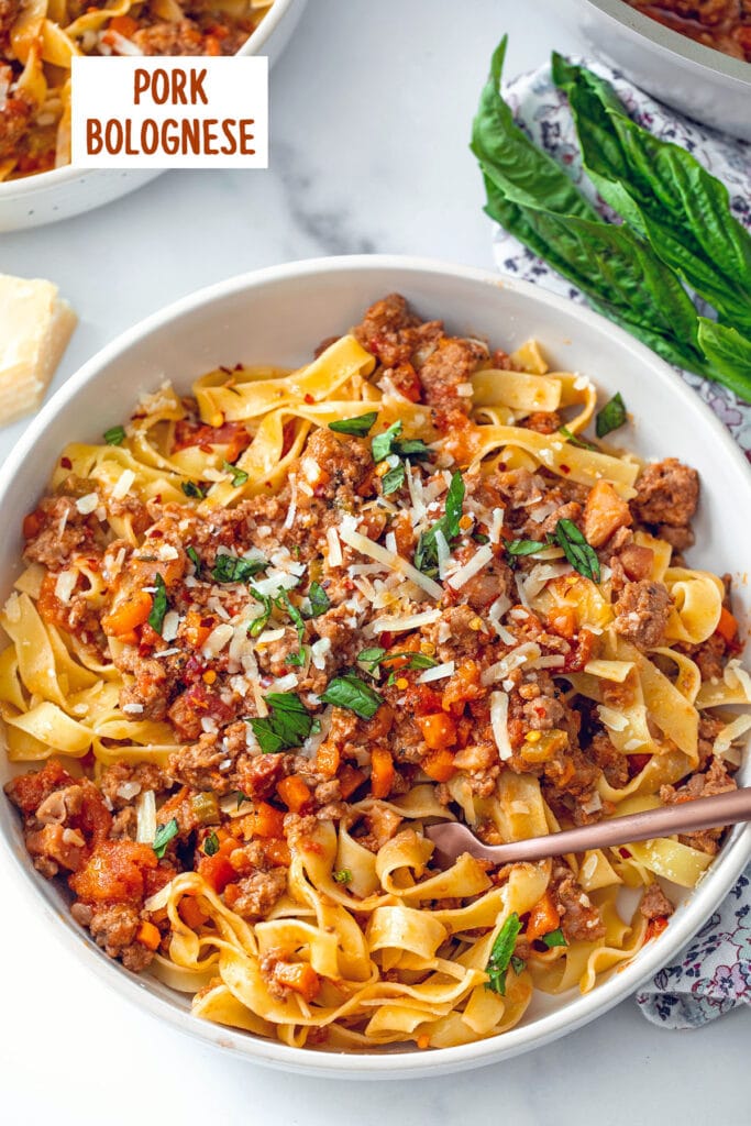 Overhead view of pork bolognese with pasta in a bowl with basil and parmesan in background and recipe title at top.
