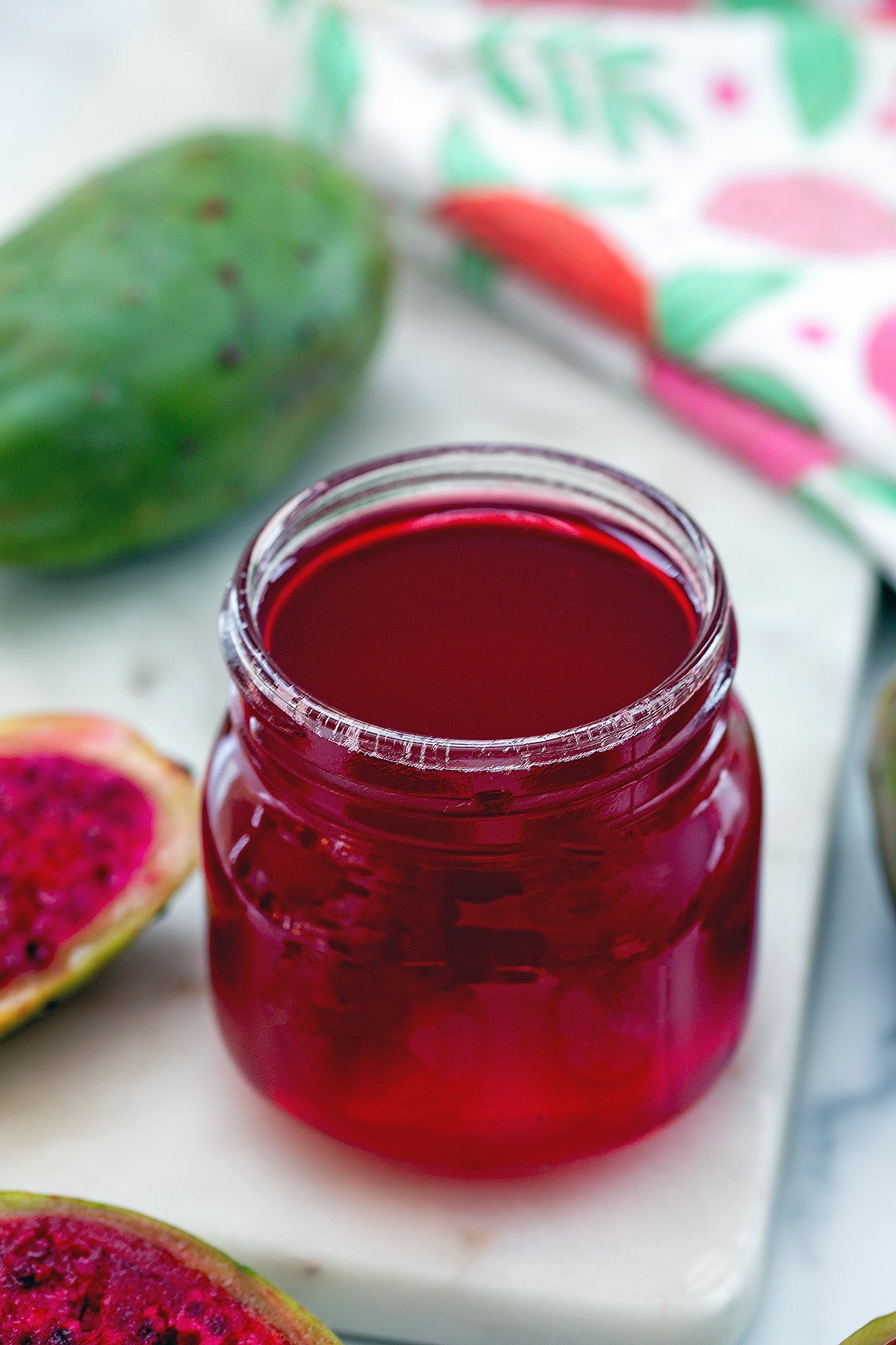 Overhead view of a small jar of pink prickly pear syrup with fruit in background.