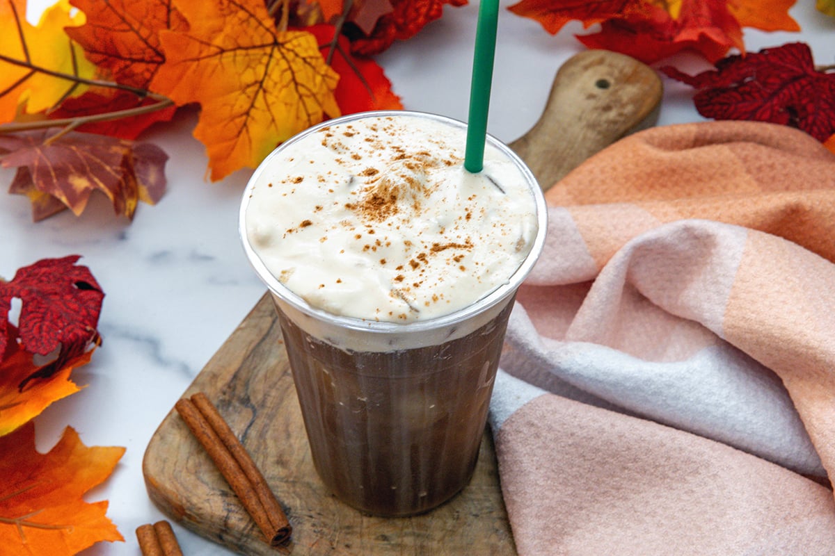 Landscape view of a cup with cold brew topped with pumpkin cold foam with pumpkin pie spice and a green straw with fall leaves all around.
