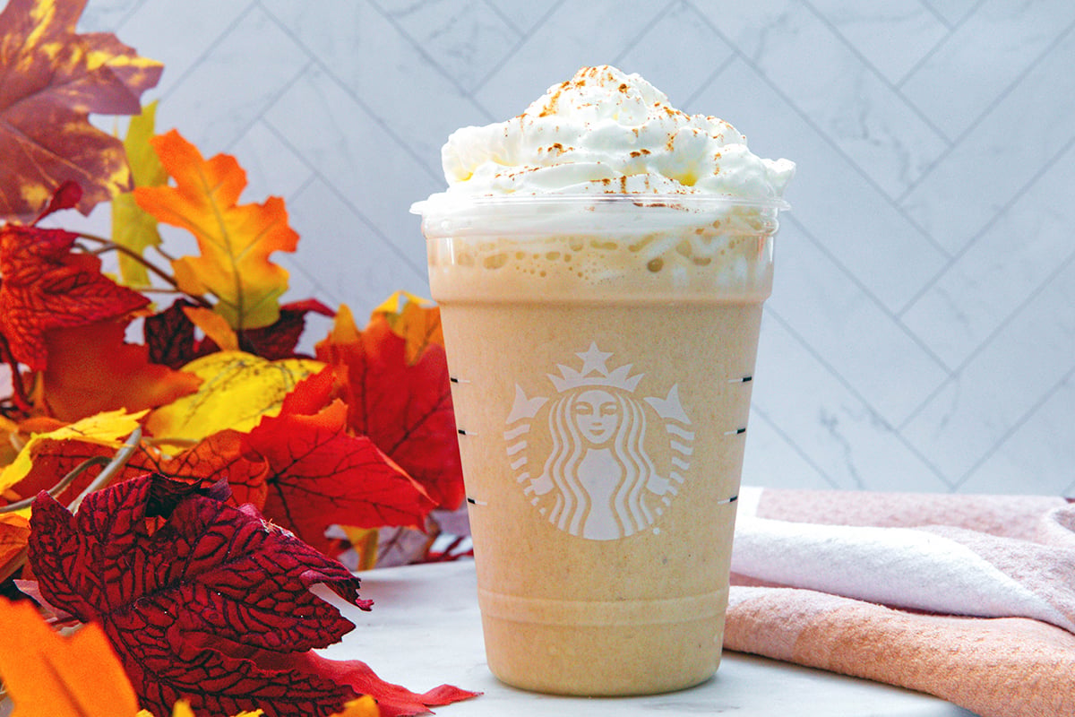 Landscape head-on view of a Pumpkin Spice Frappuccino in Starbucks cup with fall leaves in background.