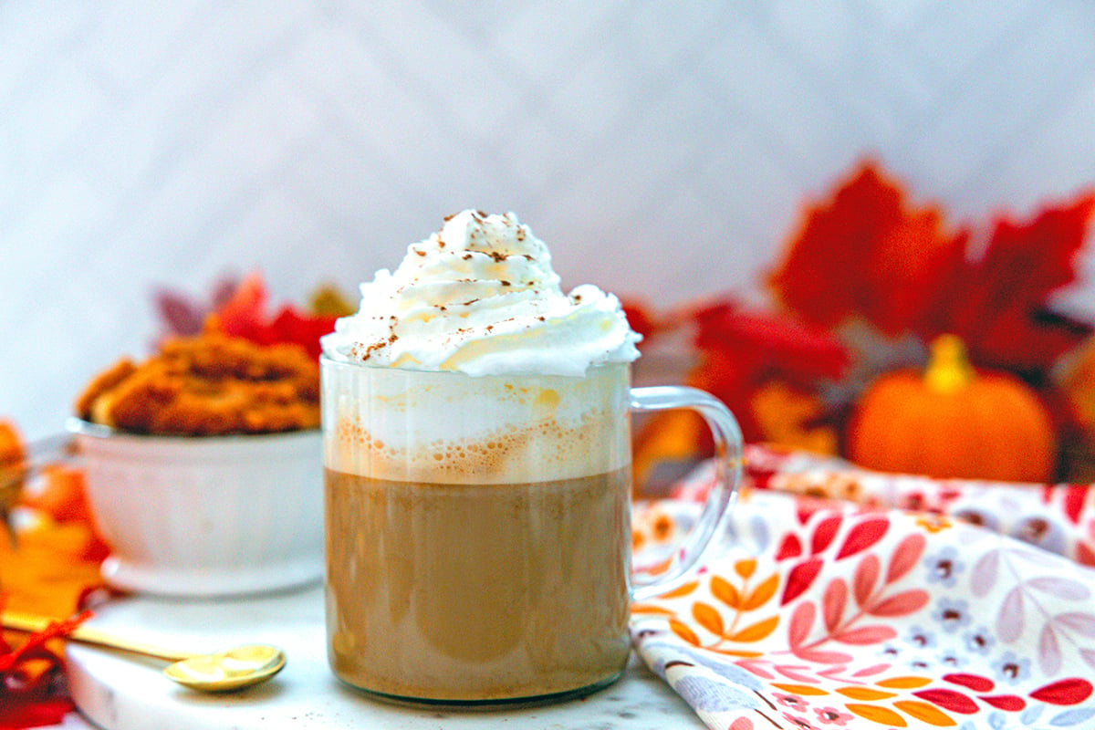 Head-on landscape view of pumpkin spice latte with whipped cream on top, heart-shaped spoon on side, and pumpkin puree in background.