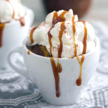 Closeup view of a pumpkin spice latte cake in a mug with whipped cream and caramel drizzle.