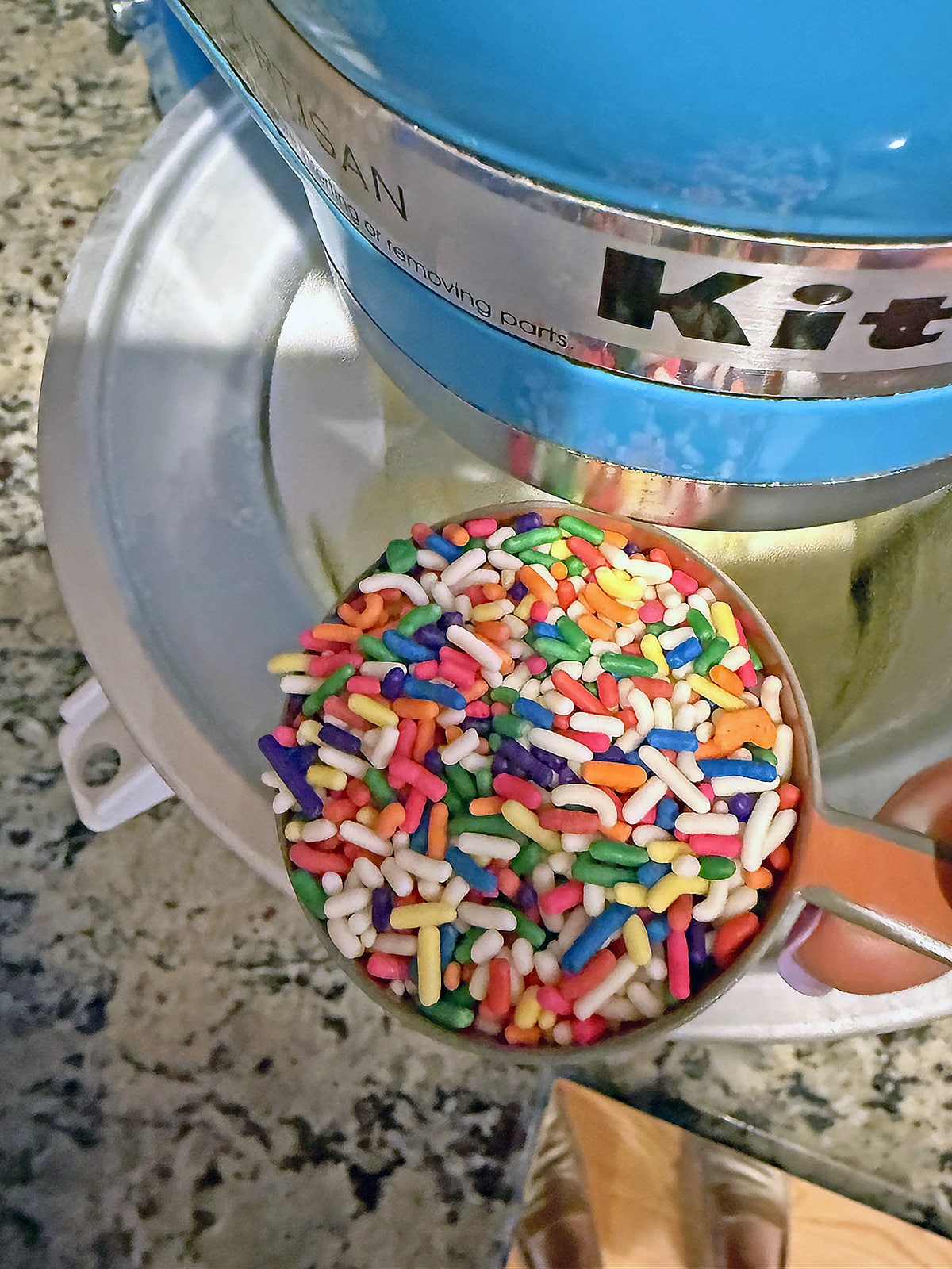 Cup of rainbow sprinkles being poured into ice cream maker.
