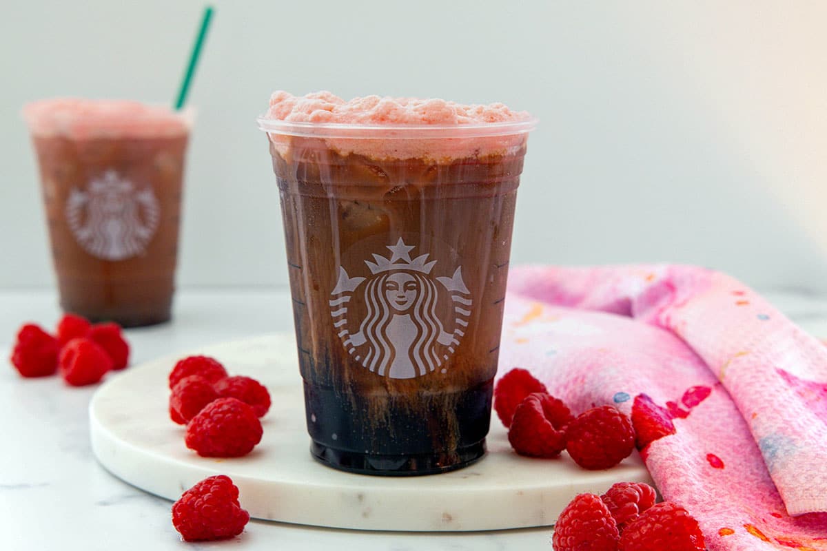 Landscape head-on view of a raspberry cream cold brew with fresh raspberries all around and second coffee in background.